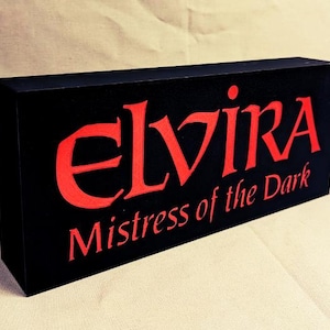 Könnte beinhalten: Schwarzes rechteckiges Schild mit dem roten Text "ELVIRA Mistress of the Dark"