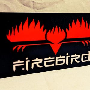 Könnte beinhalten: Schwarzes Schild mit einem roten stilisierten Vogel mit Flammen an den Flügeln. Der Text "FIREBIRD" steht in weißen Buchstaben unter dem Vogel.