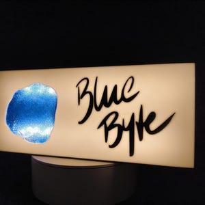 Könnte beinhalten: Ein rechteckiges, beleuchtetes Schild mit dem Schriftzug "Blue Byte" in schwarzer Schrift. Links befindet sich eine strukturierte blaue Form. Das Schild steht auf einem weißen Sockel vor dunklem Hintergrund.