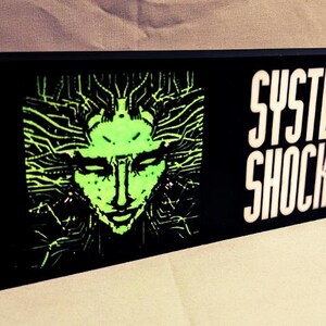 Könnte beinhalten: Ein schwarzes Leuchtenschild mit einem grünen Bild eines Gesichts, das von Schaltkreisen umgeben ist, und dem Text "SYSTEM SHOCK" in Weiß.