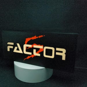 Könnte beinhalten: Ein schwarzes rechteckiges Schild mit dem Wort "FACTOR" in weißen, beleuchteten Buchstaben. Die Zahl "2" ist orange, mit einem orangefarbenen Schrägstrich-Design. Das Schild steht auf einem weißen, zylindrischen Ständer vor dunklem Hintergrund.