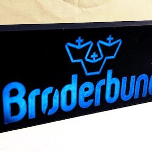 Könnte beinhalten: Ein schwarzes Schild mit dem Wort "Bruderbund" in blauer Schrift. Das Schild zeigt auch ein blaues, stilisiertes Logo mit drei Kronen.