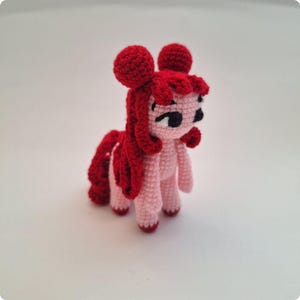 May include: A pink crocheted pony with red hair and a red tail. The pony has black eyes and a cute expression.
