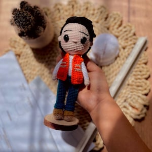 May include: A crochet doll featuring black hair, large black eyes, a white long-sleeved shirt, a bright orange vest, dark blue pants, and tan shoes. The doll stands on a small, circular wooden base and has a light-colored skin tone.