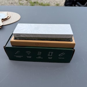 May include: A rectangular sharpening stone with a white top layer and a dark green bottom layer, resting on a light brown bamboo base. The base is set atop a dark green box labeled Royal Craft Wood, featuring icons for a whetstone, durability, bamboo, rubber, and an angle guide.