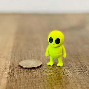 May include: A small, lime green alien figurine with large black eyes stands next to a quarter on a wooden surface. The figurine has a rounded head and a simple, two-legged body. The background is a plain white wall.
