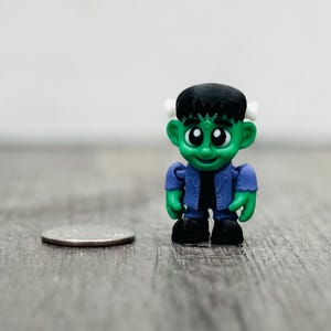 May include: A small, green Frankenstein's monster figurine with black hair, a blue jacket, and black shoes. The figurine is standing next to a coin on a wooden surface. The background is a plain white wall.