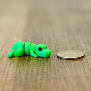 May include: Two small, bright green alligator figurines with black eyes are positioned on a wooden surface, flanking a coin. One alligator is lying down, the other is upright. The figurines are likely handmade.