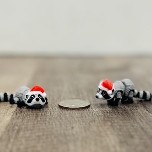 May include: Two miniature raccoon figurines, each wearing a red Santa hat, are positioned on a wooden surface. The raccoons are gray and white with black stripes on their tails. A coin is placed between them. The figurines are likely Christmas decorations.