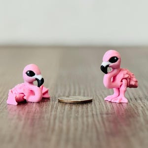 May include: Two small pink flamingo figurines with black beaks and eyes. One flamingo is lying down, the other is standing. A coin is between them. The figurines are on a wooden surface.