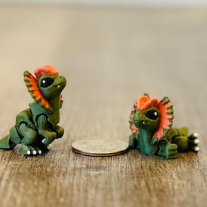 May include: Two small, handmade green and orange frilled lizard figurines. One is upright, the other is lying down. The figurines are next to a coin on a wooden surface. The lizards have black eyes and white feet.