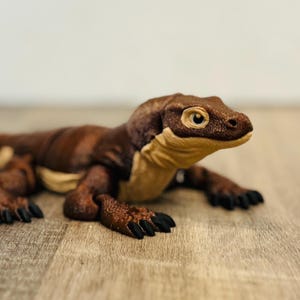 Może przedstawiać: Szczegółowa figurka smoka z Komodo, w brązowo-beżowej kolorystyce. Gad jest umieszczony na drewnianej powierzchni, prezentując swoją fakturę skóry i realistyczne cechy. Oczy smoka są jasnobrązowe.