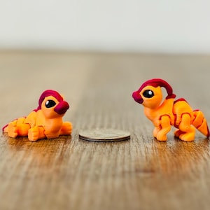 May include: Two small, orange and red dinosaur figurines. One is lying down, the other is standing. The figurines are next to a coin, suggesting their small size. The dinosaurs have a cartoonish design and are set on a wooden surface.