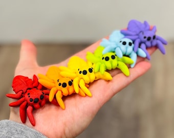 May include: A row of colorful, handcrafted spider figurines in a rainbow of hues: red, orange, yellow, green, blue, and purple. Each spider has black eyes and is posed on a hand, showcasing the vibrant color palette and whimsical design.