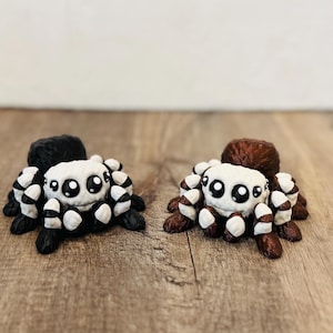 Peut inclure: Deux figurines d'araignées faites à la main. L'une est noire et blanche, l'autre est marron et blanche. Chacune a un visage blanc avec de grands yeux noirs et des pattes rayées. Les araignées sont exposées sur une surface en bois.