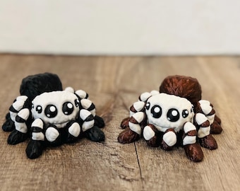 May include: Two handmade spider figurines. One is black and white, the other is brown and white. Each has a white face with large black eyes and striped legs. The spiders are displayed on a wooden surface.