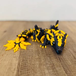 May include: A 3D-printed articulated dragon toy in black and yellow. The dragon has a detailed design with a spiky spine, a flower-like tail, and a detailed head. The toy is lying on a wooden surface.