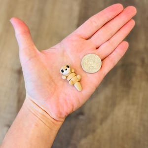 May include: A small, tan polymer clay otter figurine with black eyes and a white belly, lying on a hand next to a coin. The otter is approximately 1 inch long, with segmented body parts.