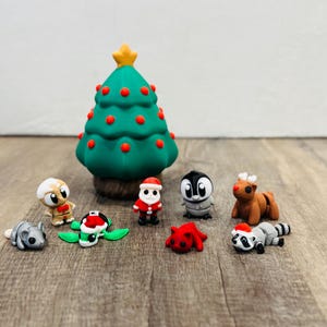 Könnte beinhalten: Eine Sammlung von Weihnachtsfiguren, darunter ein grüner Weihnachtsbaum mit roten Ornamenten und einem goldenen Stern. Weitere Figuren sind ein Weihnachtsmann, ein Pinguin, ein Rentier, ein Waschbär, eine Schildkröte und andere kleine Tierfiguren.
