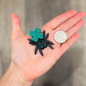 May include: A black spider figurine with a teal four-leaf clover on its back, held in a hand next to a quarter. The spider has teal accents and long legs. The clover is a bright teal color.