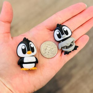 May include: Two small penguin figurines, one upright and one folded, are displayed next to a quarter. The penguins are black, white, and gray with orange beaks and yellow feet. The figurines are likely toys or collectibles.