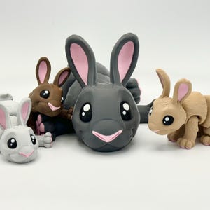 Pode incluir: Uma coleção de quatro coelhos de brinquedo em várias cores: cinza, marrom e bege. Cada coelho tem olhos grandes e expressivos e detalhes rosa nas orelhas e no nariz. Os coelhos estão em diferentes poses.