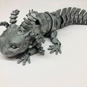 Op de afbeelding: Een grijs, metalen, gearticuleerd axolotl-beeldje met een gesegmenteerde staart. Het beeldje heeft een gedetailleerd, robotachtig ontwerp met een gestructureerd oppervlak.