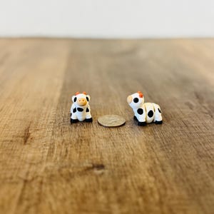 May include: Two miniature cow figurines. The cows are white with black spots, black hooves, and orange horns. One cow is standing, and the other is lying down. A coin is between the cows. The figurines are on a wooden surface.