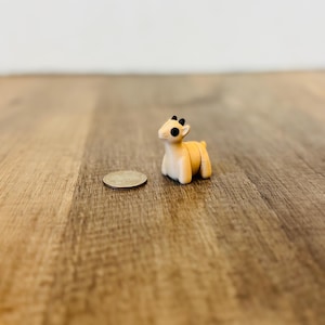 Pode incluir: Uma figura de veado em miniatura, de cor bege, com chifres e olhos pretos. A pequena escultura está colocada ao lado de uma moeda numa superfície de madeira. O veado tem aproximadamente 2,5 cm de altura.