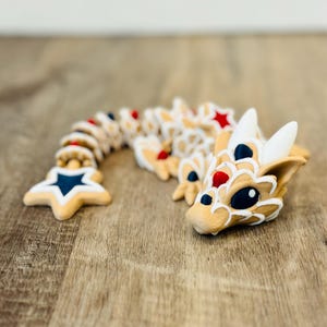 Pu&ograve; includere: Una stravagante statuetta di drago articolata in beige, bianco, rosso e blu. Il corpo del drago &egrave; segmentato, con una coda a forma di stella e una testa dettagliata. Il design presenta una combinazione di colori patriottici, con stelle e strisce.