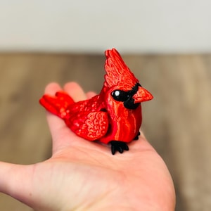 May include: A vibrant red cardinal figurine with black accents. The bird has a detailed texture, a prominent crest, and a black beak. The figurine is a decorative item, likely for display or collection.