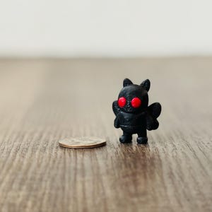 May include: A small, black figurine of a creature with red eyes, small wings, and pointed ears stands next to a quarter on a wooden surface. The figurine has a textured surface and a stylized design.