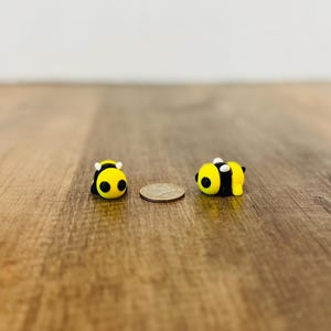 May include: Two small, yellow and black bee figurines with white accents, resting on a wooden surface. A coin is placed between the bees. The bees are in a lying down position.