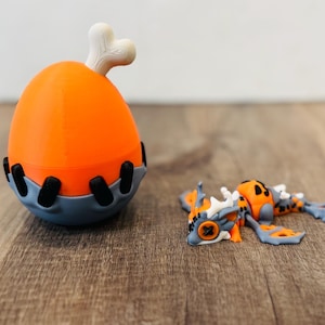 May include: An orange and gray 3D-printed toy egg with a bone on top, and a small orange, gray, and white creature. The egg has black accents and sits on a wooden surface. The creature is lying down.