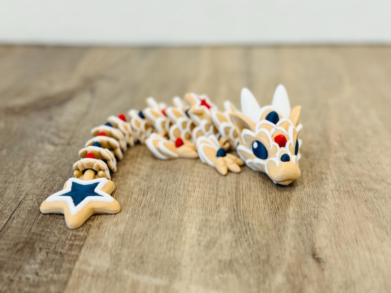 Pu&ograve; includere: Figurina di drago articolata in beige, bianco, rosso e blu, con coda a forma di stella. Il drago &egrave; posato su una superficie di legno. Il design presenta un motivo ripetuto di cerchi e una stella. Lunghezza di circa 20 cm.