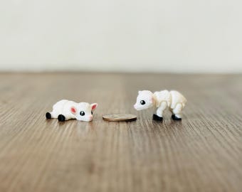May include: Two miniature white lamb figurines with black hooves and pink ears are positioned on a wooden surface. One lamb is lying down, and the other is standing. A coin is between them.