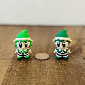 May include: Two miniature elf figurines with green hats and outfits, white trim, and large eyes. The elves are standing on a wooden surface next to a coin. The figurines are approximately 1.5 inches tall.