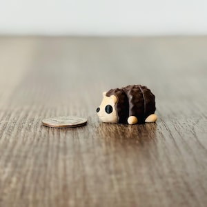 Puede incluir: Una figura de erizo en miniatura con un dorso marrón texturizado y una cara y vientre de color beige claro. La figura está junto a una moneda, sirviendo como referencia de tamaño. El erizo tiene ojos negros y pies pequeños y redondeados.