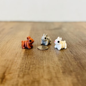May include: Three miniature bunny figurines in shades of orange, gray, and white with black eyes and a coin. The bunnies are positioned on a wooden surface, with a plain white background.