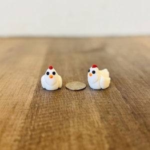 May include: Two small, white chicken figurines with red combs and black eyes sit on a wooden surface. A coin is placed between them for scale. The figurines are likely handmade and are a decorative item.