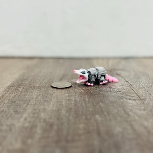 Puede incluir: Figura de zarigüeya en miniatura con pelaje gris, patas negras y cola y boca rosas. La figura está junto a una moneda, mostrando su pequeño tamaño. La zarigüeya tiene la boca abierta, como si sisease.