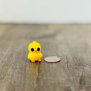 Può includere: Una piccola statuina a forma di pulcino giallo con grandi occhi neri e piedini arancioni. Il pulcino è accanto a una moneta su una superficie di legno. La statuina ha un design dettagliato e un aspetto allegro.