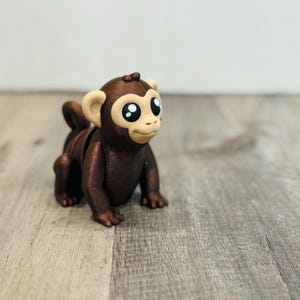 May include: A brown 3D-printed monkey figurine with large, expressive eyes and tan ears. The monkey is in a crawling position, with its arms and legs extended. The figurine is on a wooden surface.