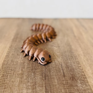 Könnte beinhalten: Eine braune, segmentierte Raupenfigur mit schwarzen Augen und kleinen Beinen. Das Spielzeug besteht aus einem festen Material und ist auf einer Holzoberfläche platziert. Ein skurriles Dekorationsstück.