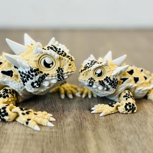 Könnte beinhalten: Zwei detaillierte Spielzeug-Krötenechsen. Die Echsen sind gelb mit schwarzen Flecken und weißen Stacheln. Sie haben große, runde Augen und sind einander zugewandt. Der Hintergrund ist eine Holzoberfläche.
