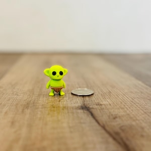 May include: A small, lime green figurine of a cartoon alien with large black eyes and a brown diaper. A silver coin is next to the figurine. The figurine is approximately 1.5 inches tall.