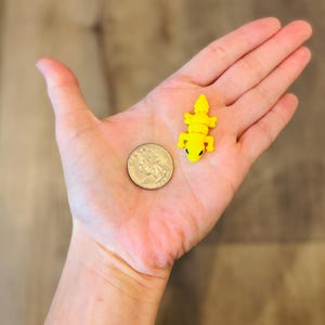 May include: A yellow articulated lizard toy with black eyes is held in a person's hand next to a quarter. The toy is small, likely a miniature figurine or charm. The quarter provides a size reference.
