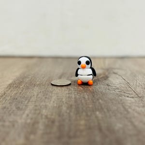 May include: A miniature penguin figurine with a black head, white body, and orange feet and beak. The penguin is next to a quarter coin for size comparison. The figurine is on a wooden surface.