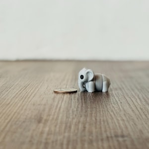 May include: A small, gray elephant figurine with black eyes stands next to a coin on a wooden surface. The elephant is a miniature sculpture, likely a decorative item or toy. The coin provides a size reference.