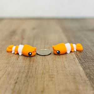 Pode incluir: Duas figuras de peixe-palhaço laranja e branco com olhos pretos estão posicionadas sobre uma superfície de madeira. As figuras estão ao lado de uma moeda. O fundo é uma parede de cor clara.